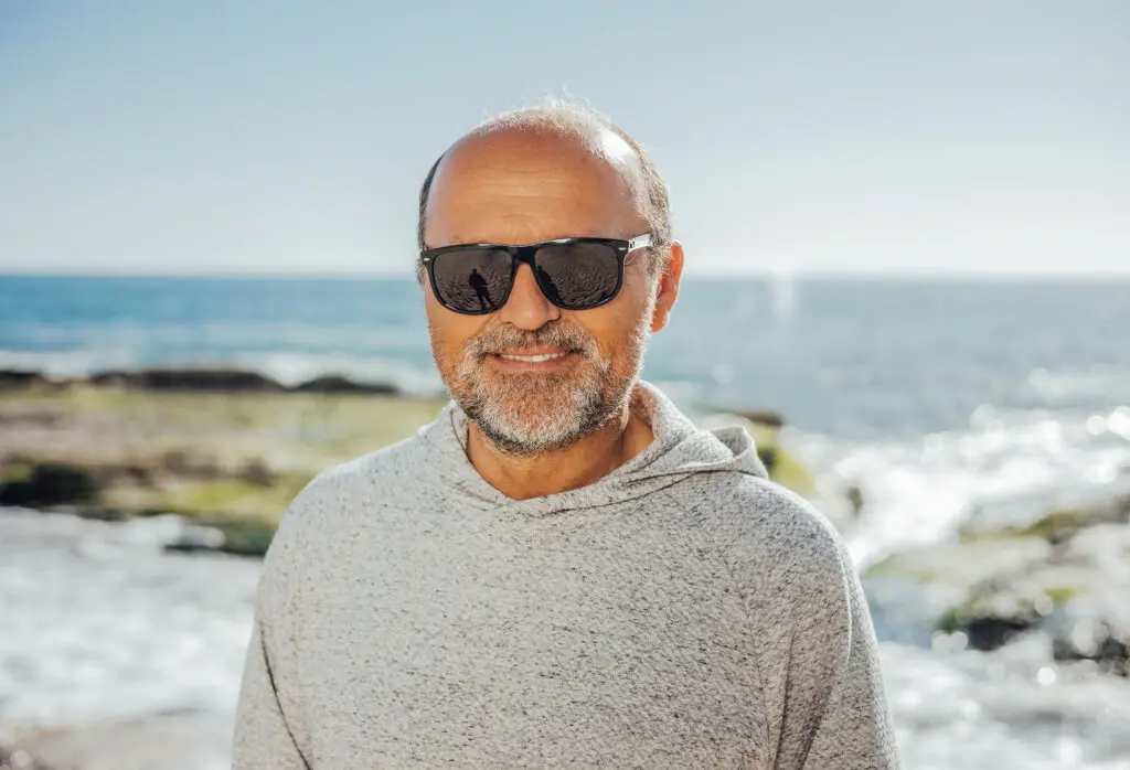 An elderly man smiling and wearing sunglasses on a rocky beach