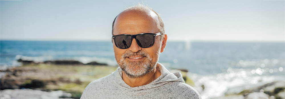 An elderly man smiling and wearing sunglasses on a rocky beach